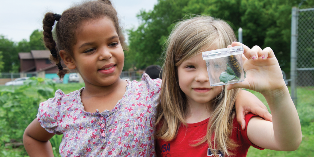 Photo of two girls outside looking at a caterpillar in a container.