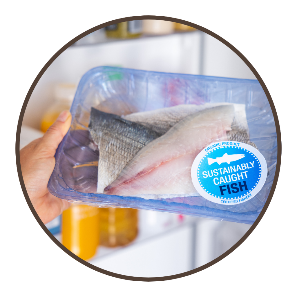 A close up of a hand holding a package of raw fish with a sticker that says, "sustainably caught."