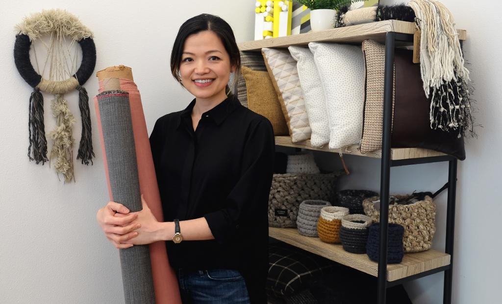 Aileen smiles and holds bolts of fabrics in front of a display of REVISION GOODS products.