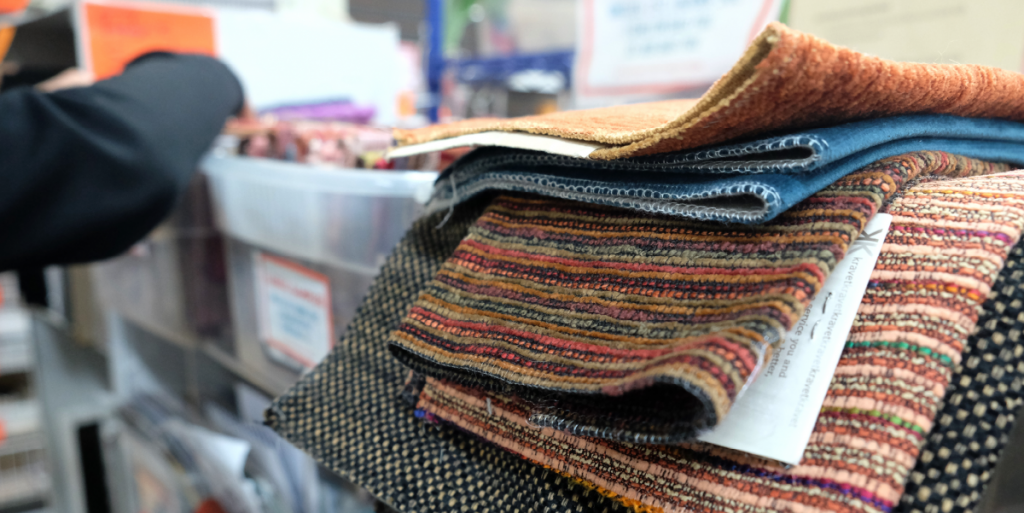 A pile of fabric sits on a container. In the background, Aileen searches through fabric swatches.