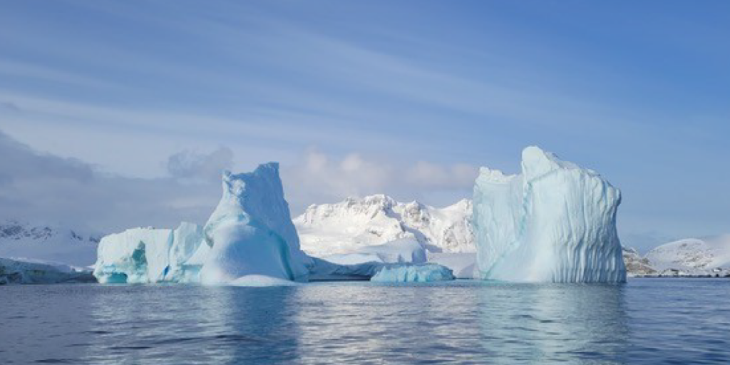 A pale blue iceberg sits in the water. Blue skies rise behind it.