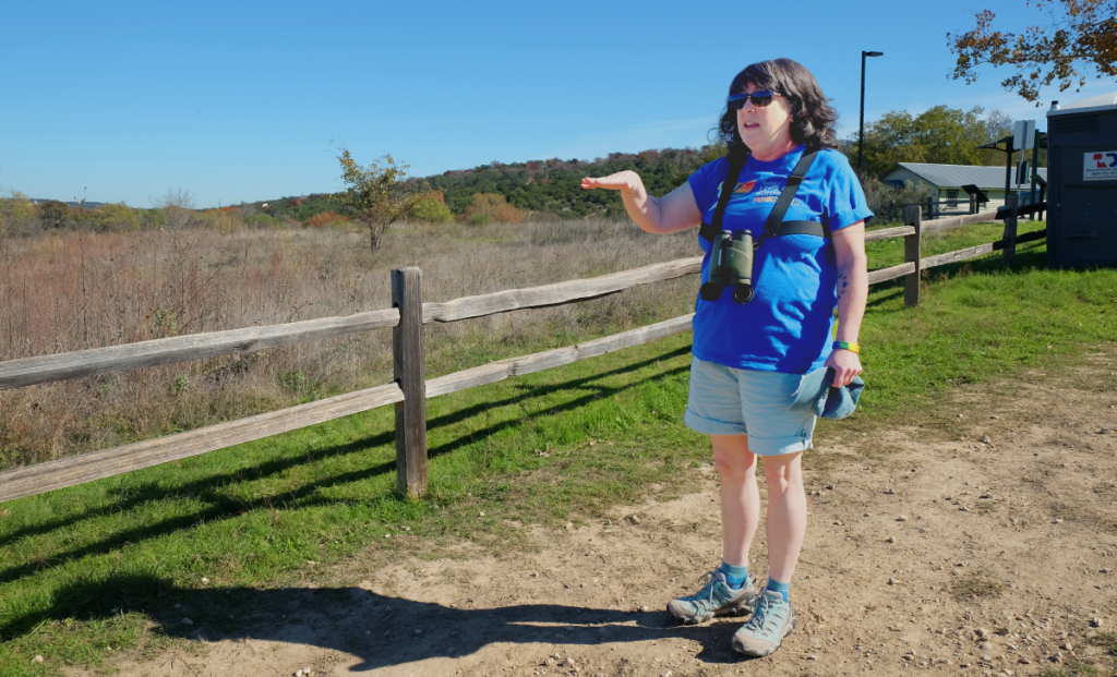 Amy gestures to the land while standing on a path at Commons Ford Ranch. Binoculars lay around her neck.