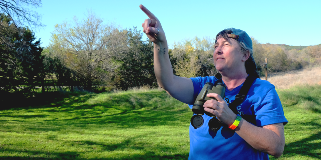 Amy points in the distance. She is holding binoculars and wearing a rainbow bracelet and backwards baseball hat.