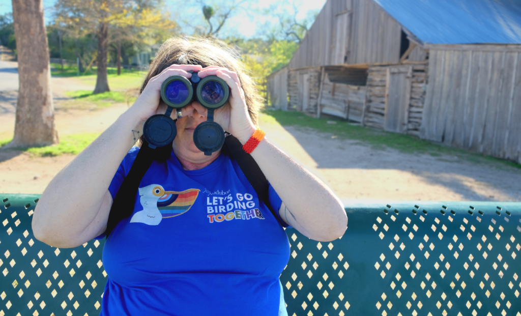Amy sits on a bench with binoculars held to her eyes. Her shirt has a pelican with the pride flag in its beak and reads, "Audubon: Let's Go Birding Together."