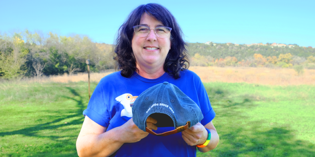 Amy smiles and holds a baseball hat that reads, "bird nerds since 1952."