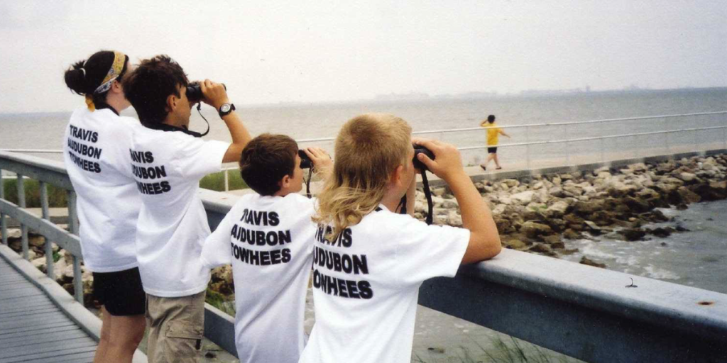 Kids stand at a pier overlooking the water. Each holds binoculars up to their eyes and wears a white tee shirt that says, "Travis Audubon Towhees."