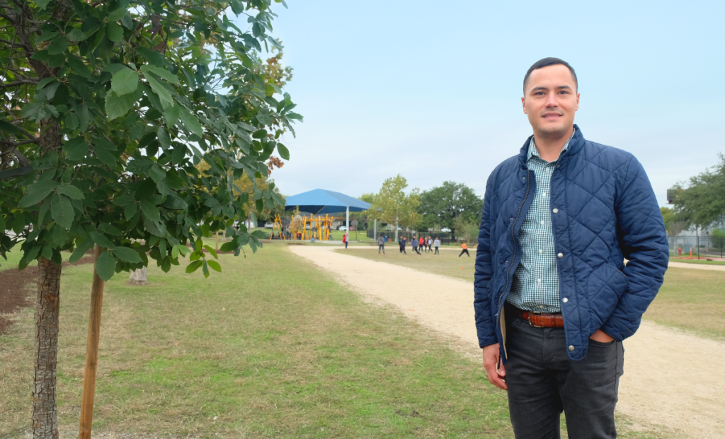 Andrew stands next to a medium-sized planted tree. Kids run around on the campus behind him.