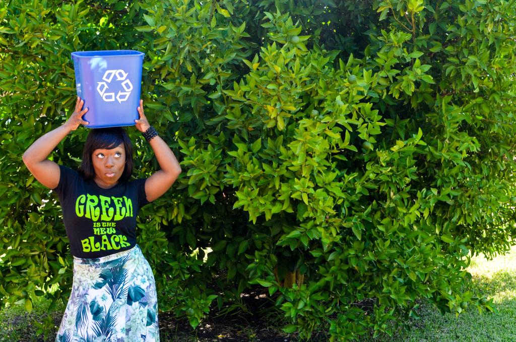 CarShandra Hollins in front of a tree wearing a "Green is the New Black" shirt and holding a recycling bin on her head.