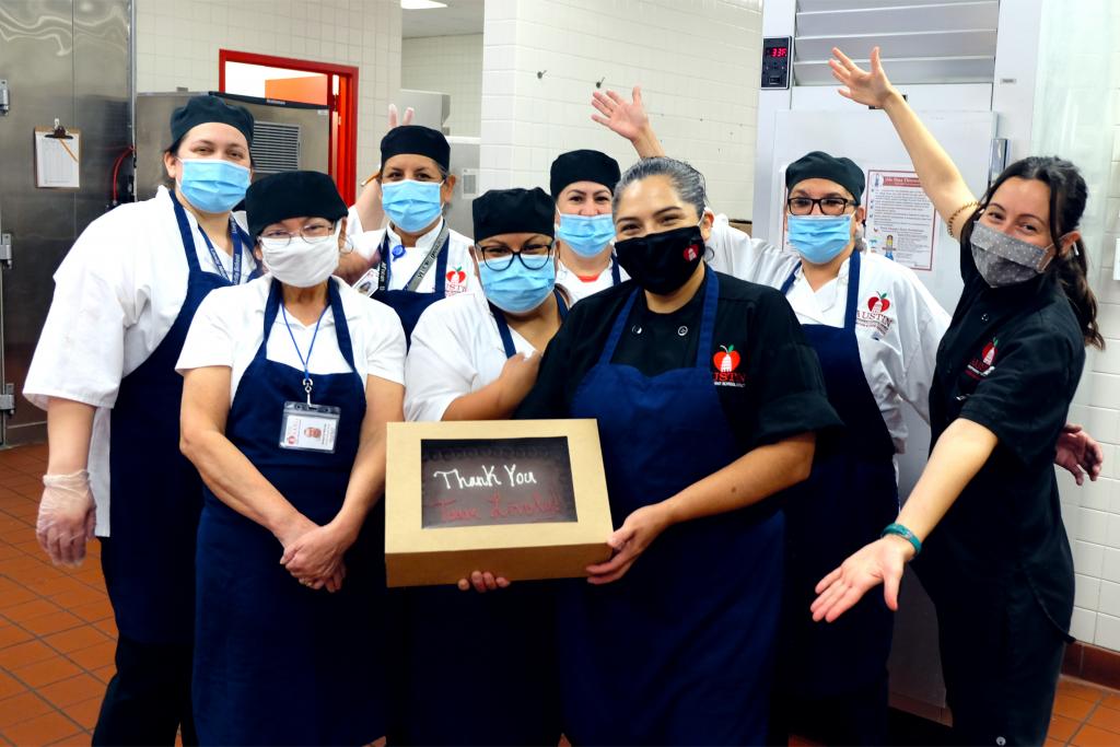A photo of the kitchen team at Lively Middle School. The manager in the middle holds a cake. Diane stands to the right with her arms wide open.