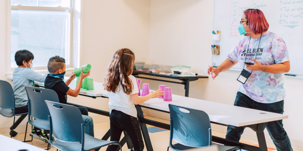 A person stands at a whiteboard. Young students work on an activity at a row of desks in front of them.