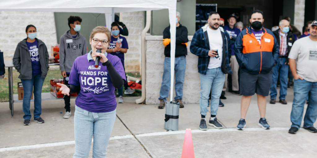 Dr. Murillo speaks into a microphone outside. Around her, people attentively listen.