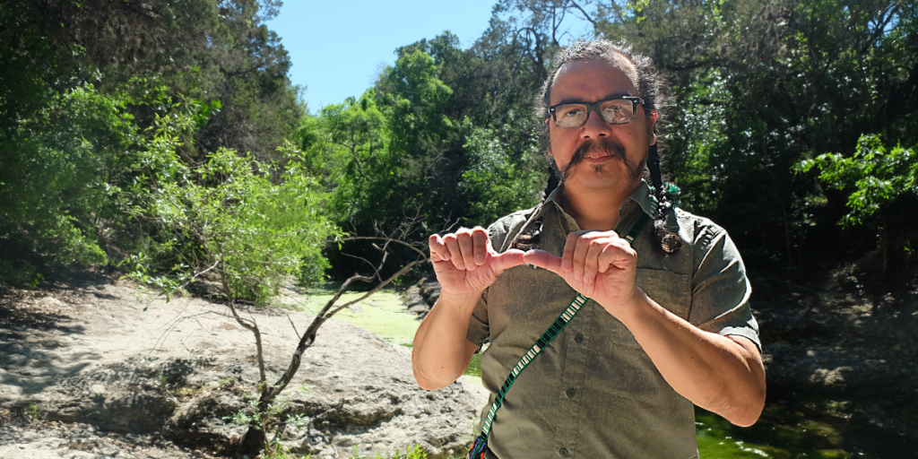 Gabriel stands in front of Blunn creek. He holds the tips of his two thumbs together with his fingers folded towards his palms.