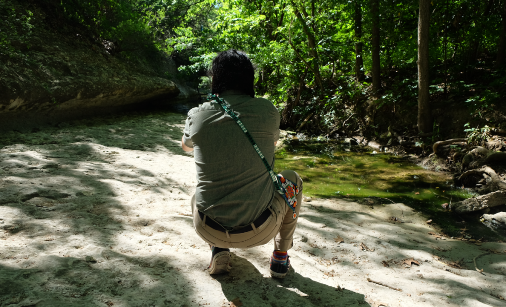 Gabriel, photographed from behind, squats with his camera phone looking out to Blunn Creek.