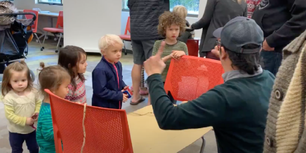 Young students gather around a table that Gabriel is sitting at. He is signing to them.