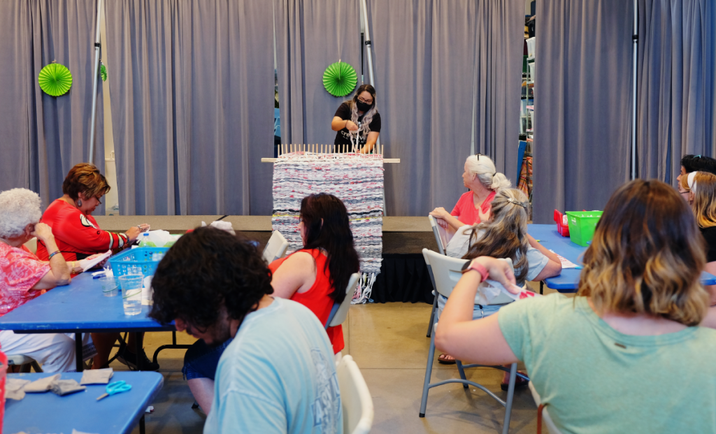 Kiana stands on a stage at the front of a large room. People at tables looking up at her as she demonstrates how to use a wooden peg loom.