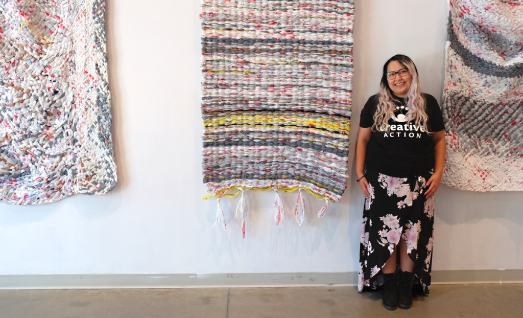 Kiana stands in the lobby of the UpFront Gallery with hanging woven mats behind her.