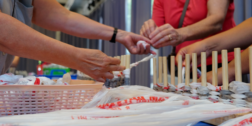 Three sets of hands work on a wooden hand loom with balls of plarn.