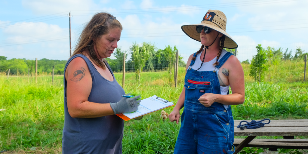 Leatha checks items off a list on a clipboard while talking to a woman in a large sunhat.