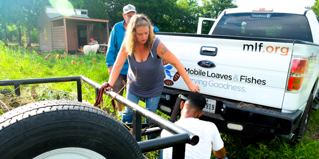 Leatha stands behind a pickup truck, trying to attach a trailer. Two other people are helping her.