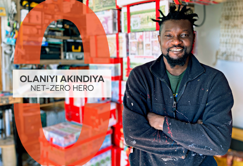 A photo of Olaniyi Akindiya standing in their studio smiling. Behind him is an artwork in progress using recycled cardboard boxes. A graphic on it reads, "Olaniyi Akindiya: Net-Zero Hero".