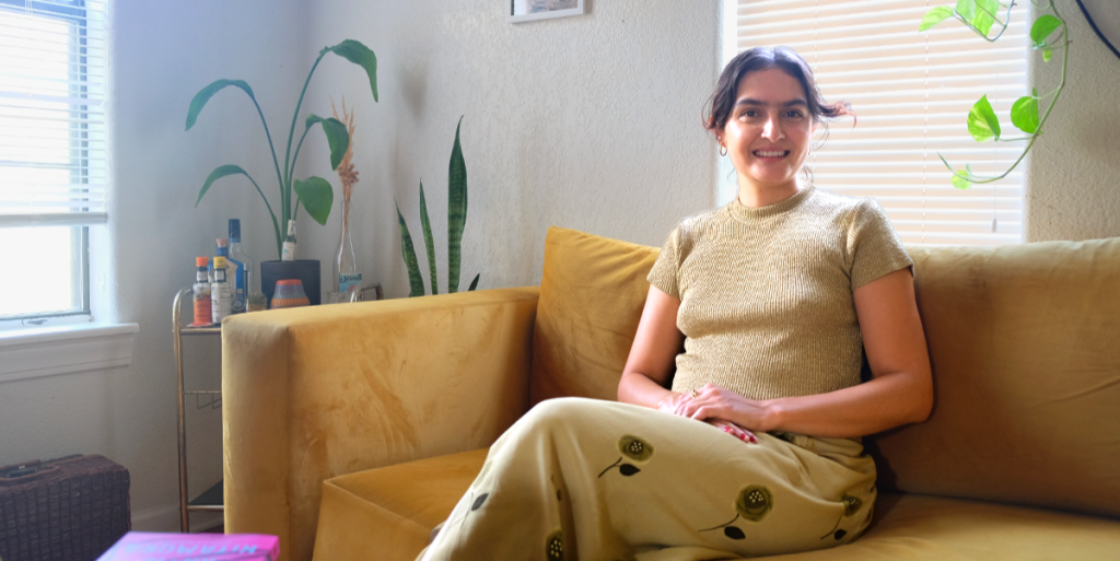 Reza smiles while sitting on her couch. Plants are all around her.