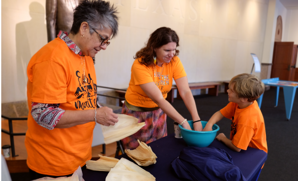 Two women stand behind a table. A young boy stands in front of them. One woman pulls corn husks apart. The other woman and boy have their hands in a large bowl of water.