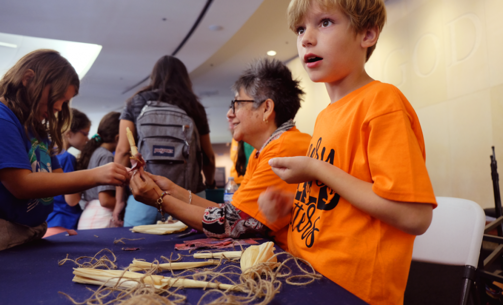 A boy talks animatedly behind a table scattered with corn husk dolls.