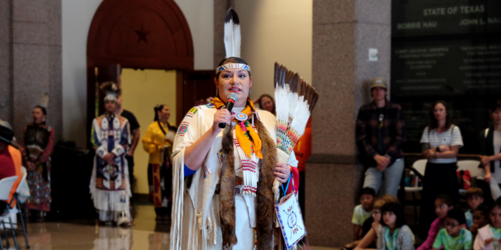 A woman dressed in an Indigenous dress speaks into a microphone.