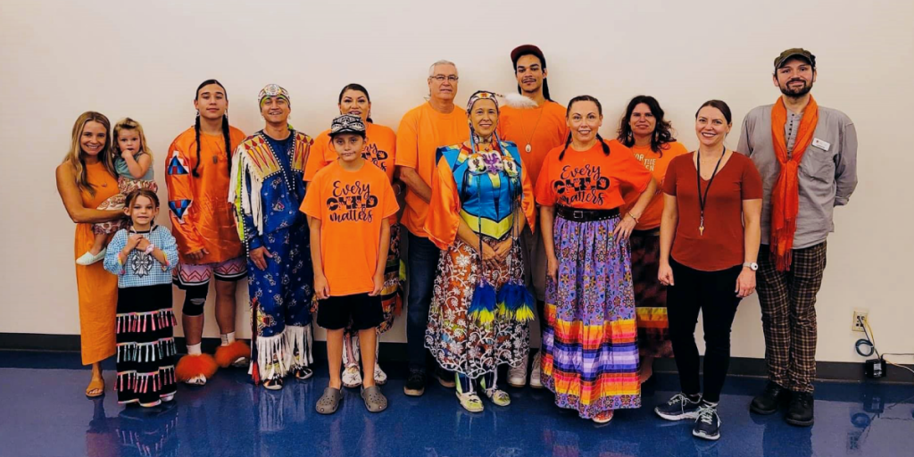 A group of people stand together smiling. Many of them are wearing orange shirts. Some are dressed in Indigenous clothing.