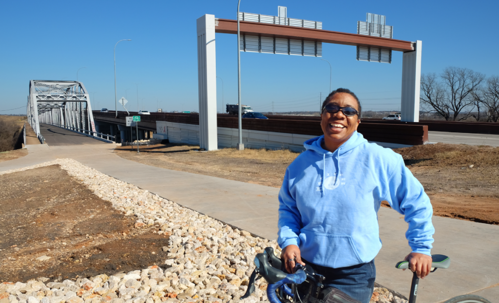 Stephanie smiles next to her bike. Montoplis Bridge and Ed Bluestein Blvd. are seen behind her.