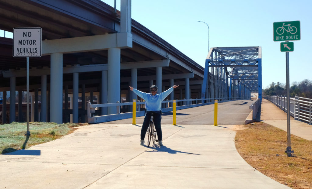 Stephanie stands on her bicycle in front of Montoplis Bridge with both hands in the air.