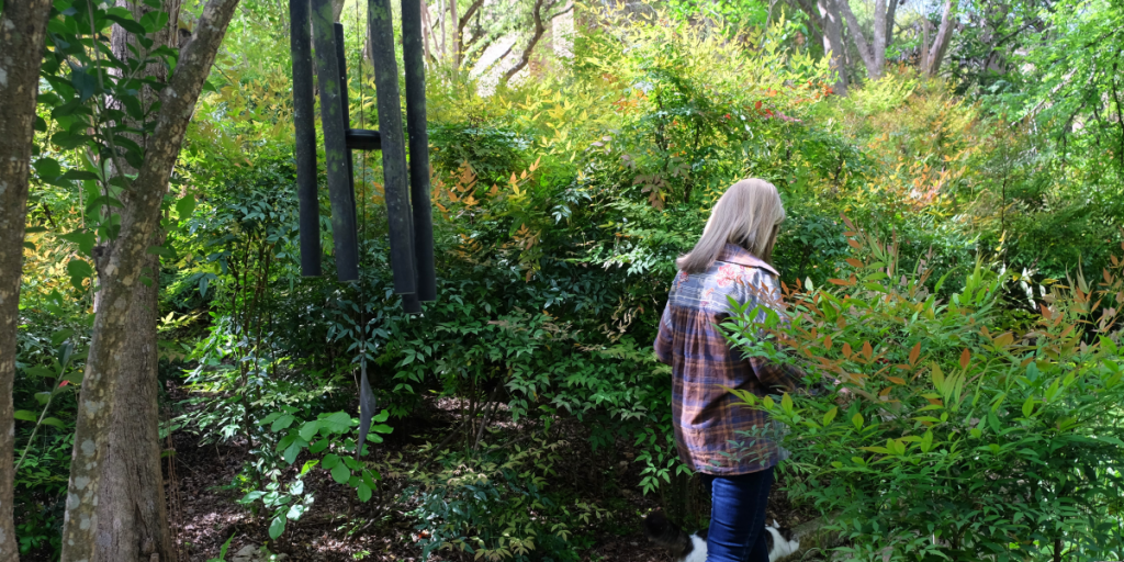 Susan walks through a bushy area. A windchime is to her left.