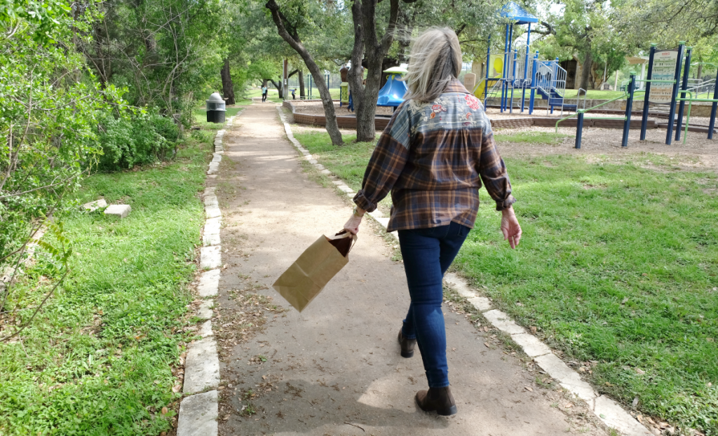 Susan walks down a path away from the camera. A paper bag is in her hand.