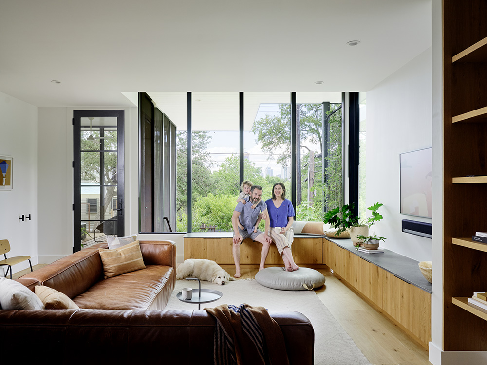 Trey and his family sit on a bench in their home. Large windows looking out to trees are behind them.