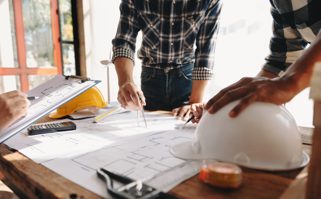 men pointing at blueprints on a table with a hardhat