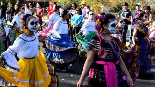 A group of dancers walking down a street during a parade. They are dressed to celebrate the Day of the Dead, or Dia de los Muertos, with flowers in their hair, colorful clothing, and sugar-skull face paint.