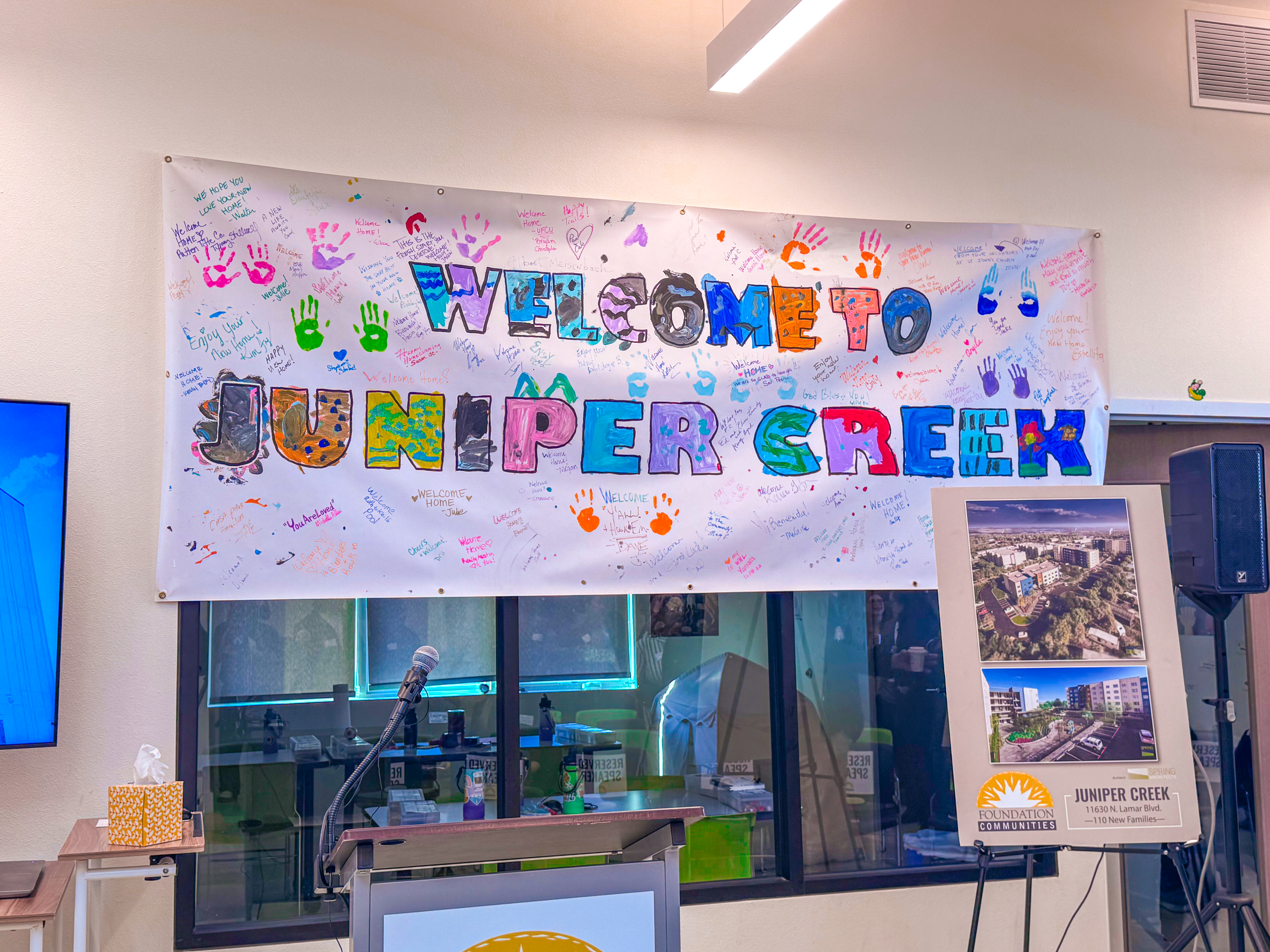 “Welcome to Juniper Creek” banner decorated with colorful handprints and handwritten messages from residents, displayed above a podium and microphone during the Juniper Creek grand opening event. An easel sign shows renderings of the 110-unit affordable housing development.
