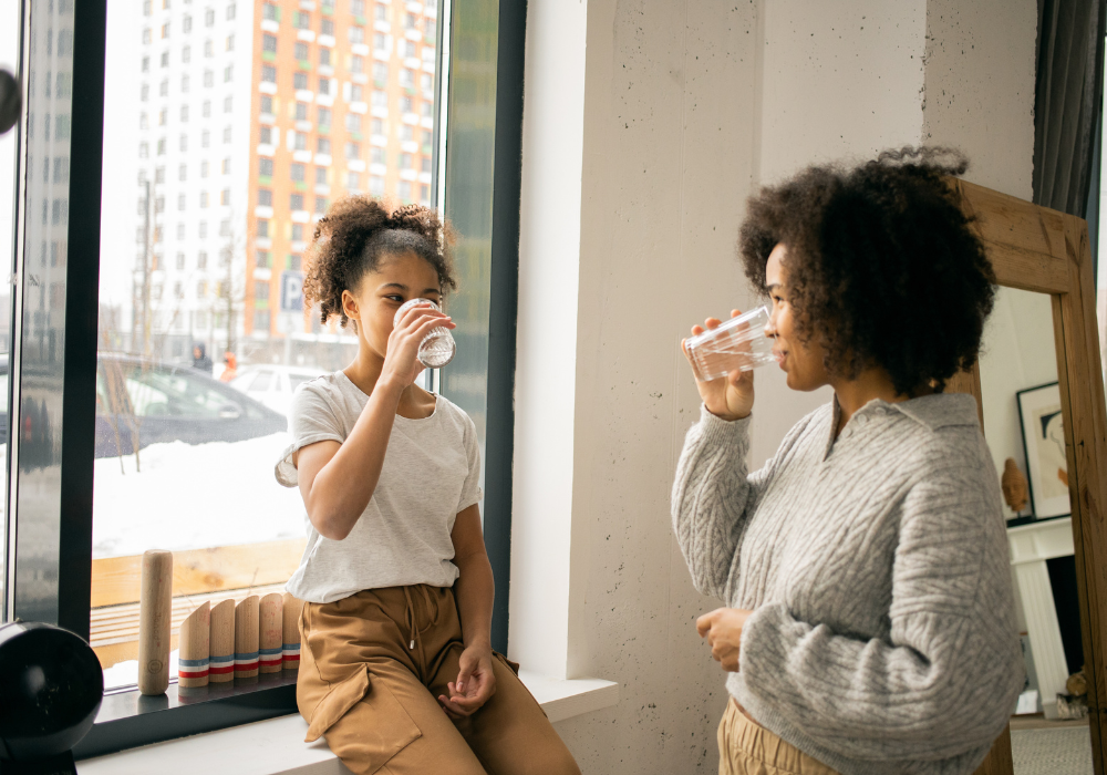 A woman and her daughter drinking a glass of water by a window.