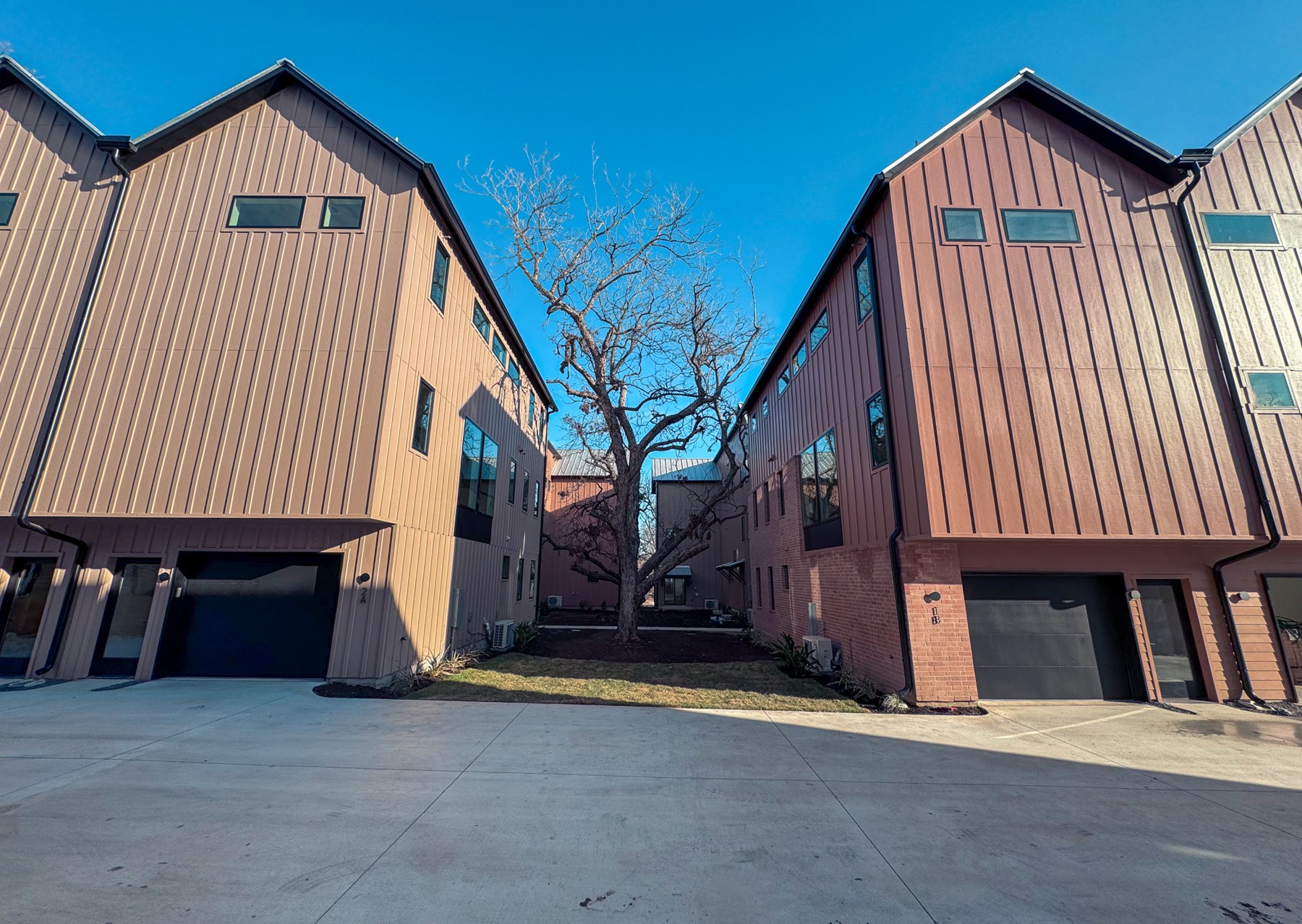 Two modern townhouse buildings face each other across a concrete driveway, with a leafless tree centered between them under a clear blue sky.