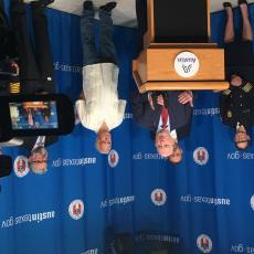 Middle-aged man in blue suit stands behind podium speaking at a press conference. Other government officials stand behind him in front of a curtain with the City of Austin seal and logo on it.