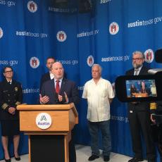 Middle-aged man in blue suit stands behind podium speaking at a press conference. Other government officials stand behind him in front of a curtain with the City of Austin seal and logo on it.