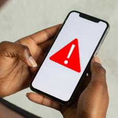 image of an African-American woman's hands holding a cell phone. The phone has an image of a red triangle and exclamation mark to indicate an alert.