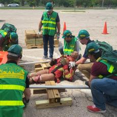 A group of individuals wearing Community Emergency Response Team vests and personal protective gear including gloves, helmets, and goggles use wood and pipes as "cribbing" to create a lever and lift a heavy object off of a dummy in a simulated emergency response.