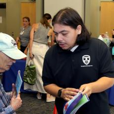 A photo showing a HSEM staff member sharing preparedness information with a community member, while people visit preparedness booths in the background.