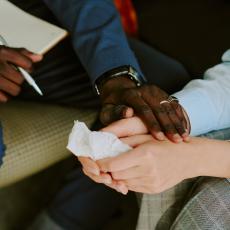 tight photo of a black man's and white woman's hands. The man holds a notepad and pen in one hand and holds the woman's hands with the other. She holds a facial tissue.
