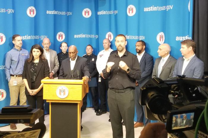 image of black man speaking behind podium at a press conference. A group of city officials stand behind him and an ASL interpreter signs next to him.