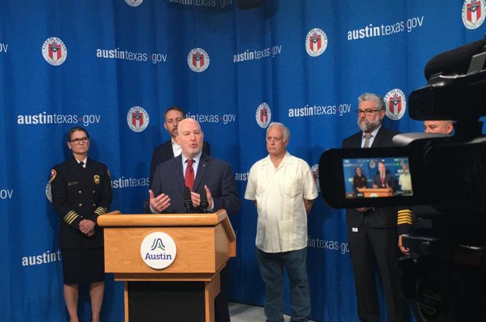 Middle-aged man in blue suit stands behind podium speaking at a press conference. Other government officials stand behind him in front of a curtain with the City of Austin seal and logo on it.