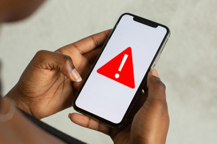 image of an African-American woman's hands holding a cell phone. The phone has an image of a red triangle and exclamation mark to indicate an alert.