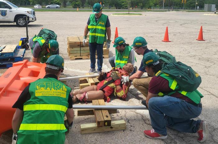 A group of individuals wearing Community Emergency Response Team vests and personal protective gear including gloves, helmets, and goggles use wood and pipes as "cribbing" to create a lever and lift a heavy object off of a dummy in a simulated emergency response.