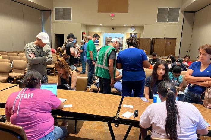 In the foreground, the image shows two women sitting at side-by-side tables with laptops, offering assistance to women who are sitting opposite of them. In the background, additional residents are sitting in rows of chairs and filling out paperwork.